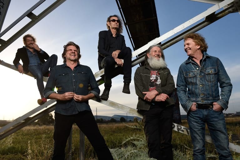 Five band members stand in casual poses on an aeroplane stairway in an open field under clear blue sky.
