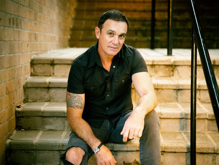 Man in black shirt crouching on brick steps against a weathered brick wall.