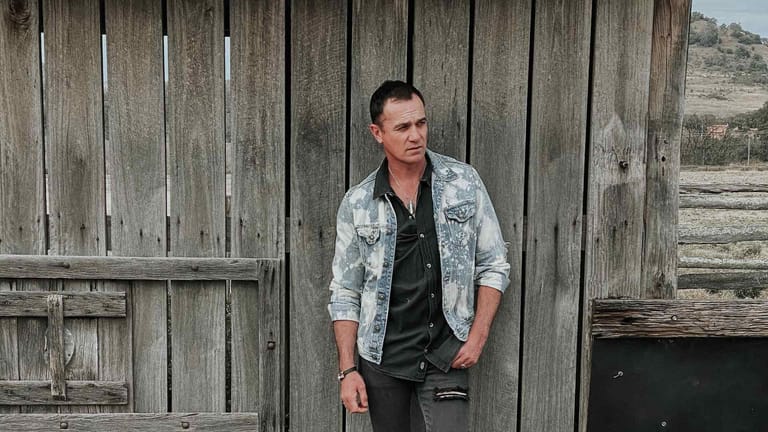 Man in a denim shirt leaning against weathered wooden barn doors in a rural setting.