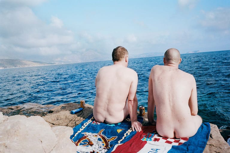 Two shirtless men sit on a rocky outcrop overlooking calm blue sea and distant coastline.