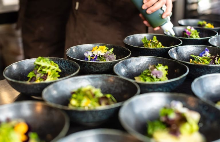 Multiple dark bowls of plated food with green garnish and edible flowers arranged for service.
