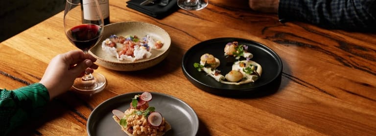 Hand reaching for wine glass among plated dishes on wooden table during dining experience.