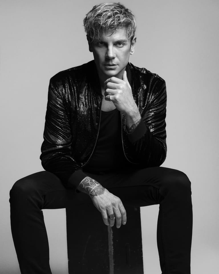 Person in black leather jacket and dark clothing seated against white background, hand to chin in contemplative pose.