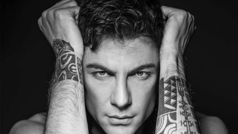 Young man with tattooed forearms posed against dark background, hands behind head.