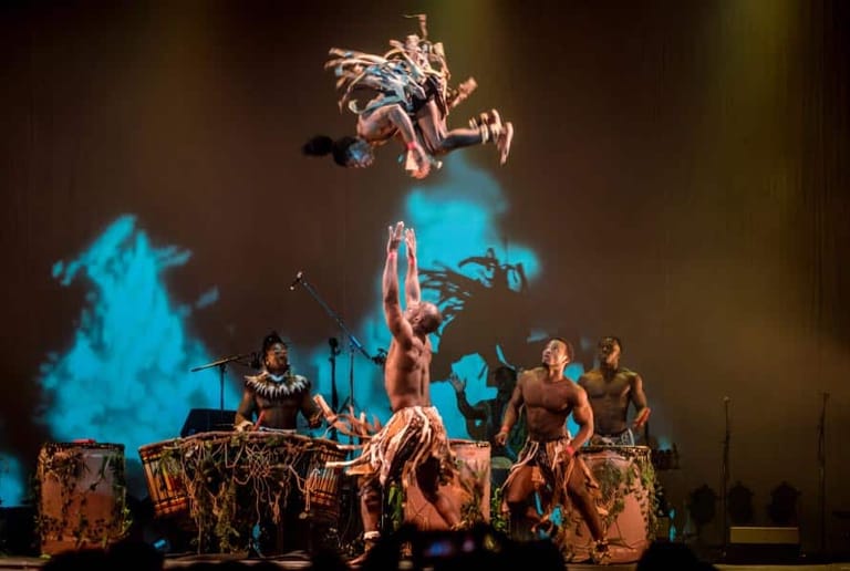 Performer leaps above a band on stage, illuminated by blue and amber lighting during a theatrical production.