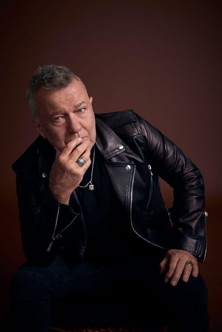 Man in black leather jacket with hand to face against warm brown background.