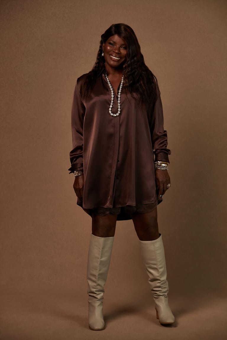 Woman in brown satin shirt and cream boots smiling against neutral tan background.
