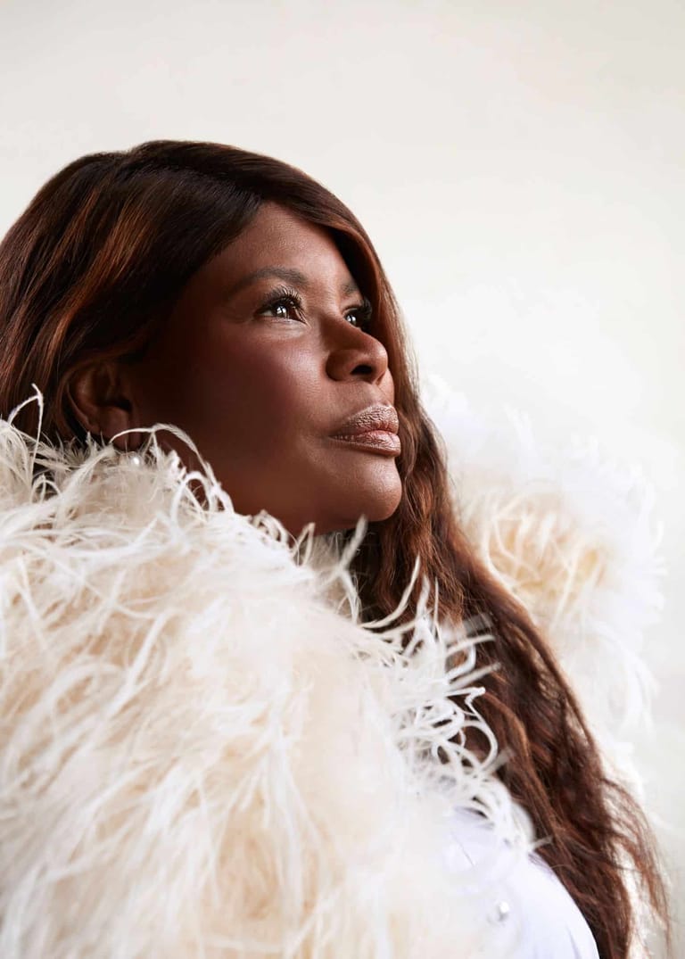 Woman with long brown hair wearing a cream feathered stole, gazing upward against a neutral background.