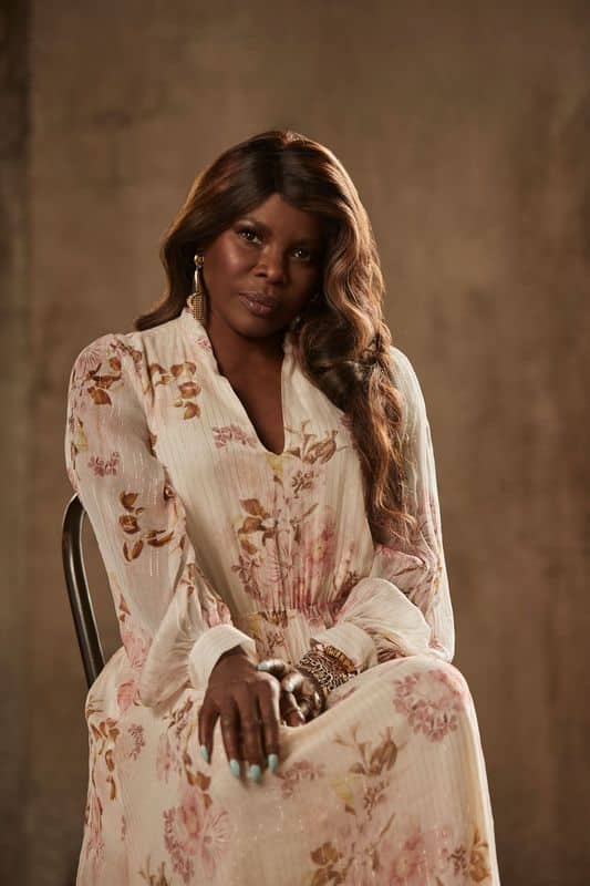 Woman in a cream floral dress seated against a neutral backdrop, gazing thoughtfully to the side.
