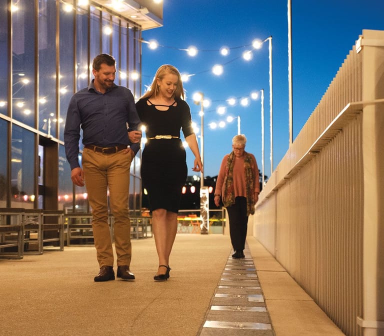 Two people walk together along a contemporary pedestrian pathway at dusk, lined with illuminated street lights.