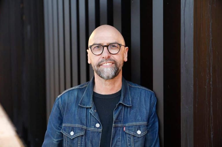 Bald man wearing glasses and a denim shirt stands against a dark corrugated metal backdrop.