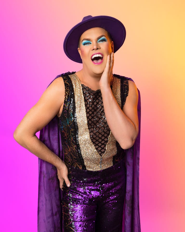 Person in sequined purple outfit and dark hat smiles against a vibrant pink-to-yellow gradient background.