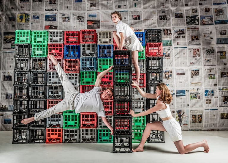 Three performers in white clothing acrobatically balance on coloured plastic crates against a newspaper-covered wall.