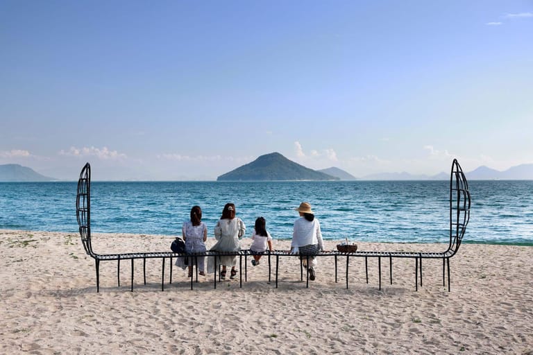 Family of four seated at a table on a beach facing a volcanic island across turquoise water.