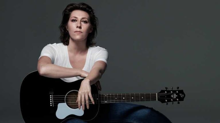 Musician with dark hair holding a black acoustic guitar against a grey background.