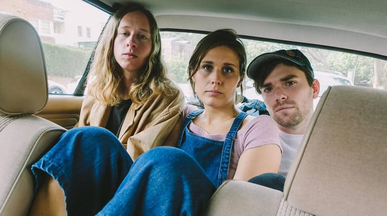 Three people sit together in the back seat of a car, looking toward the camera with serious expressions.