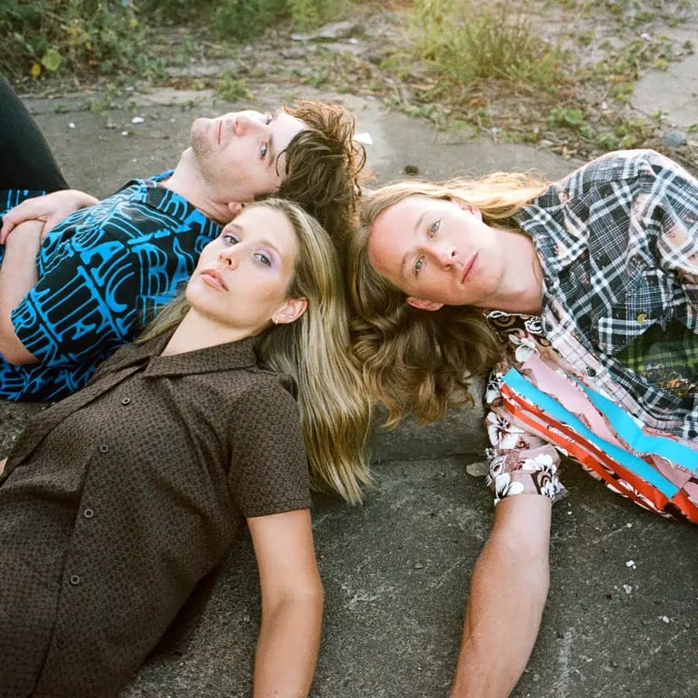 Three young people lying on pavement looking upward, wearing casual checkered and patterned shirts.