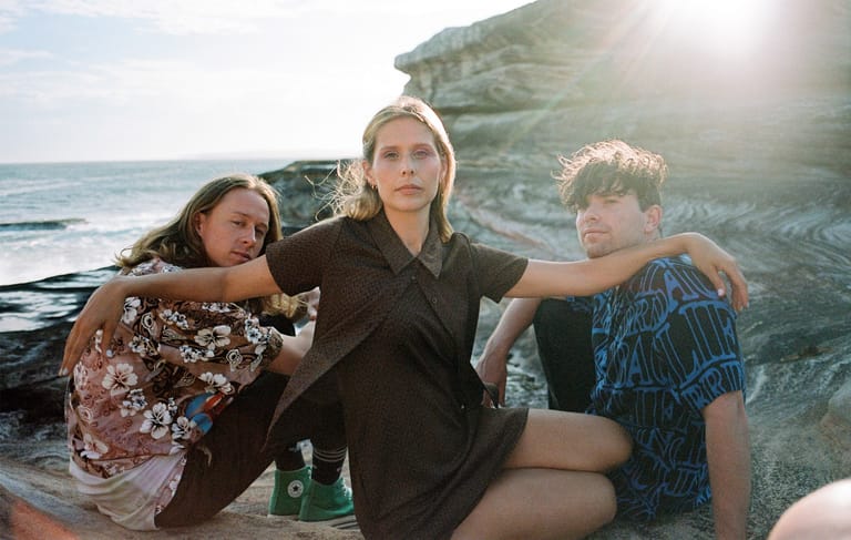Three young people pose together against a coastal cliff backdrop with ocean and sunlight visible.
