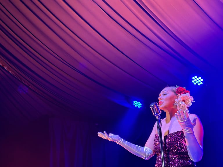 Singer in sparkly dress performing at microphone under purple stage lighting with pink floral accent.