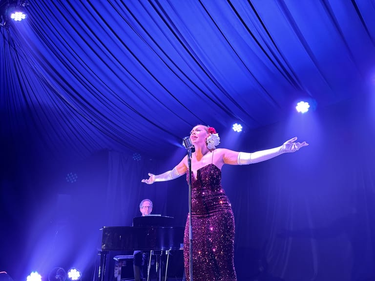 Singer in a shimmering burgundy dress performs at a microphone with arms outstretched under blue stage lighting, accompanied by a pianist.
