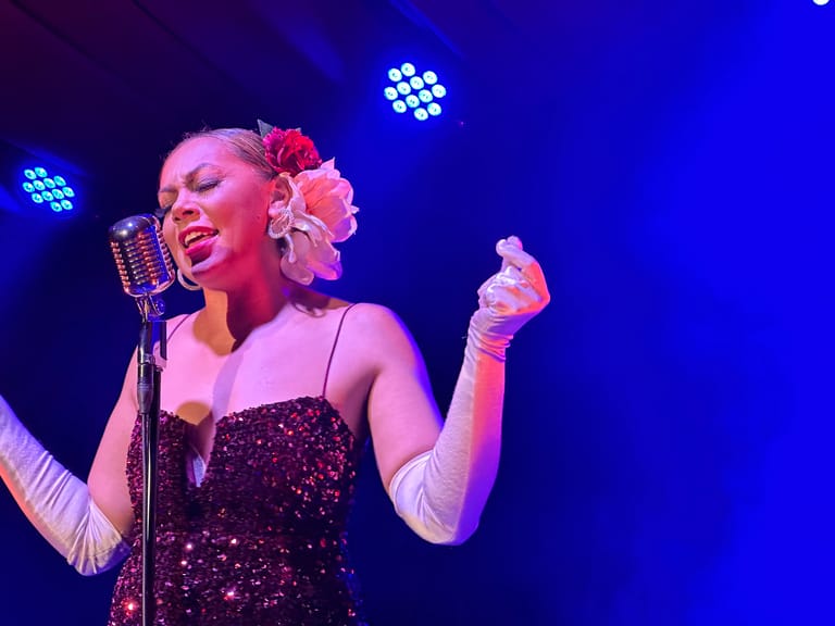 Performer in sequined dress and pink flowers sings at vintage microphone under blue stage lighting.
