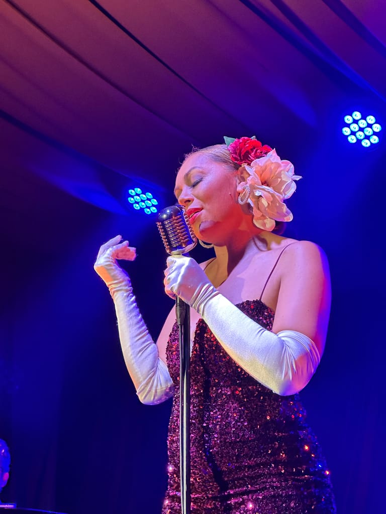 Singer in sequined dress and white gloves performs at microphone under blue stage lighting, flowers adorning her hair.