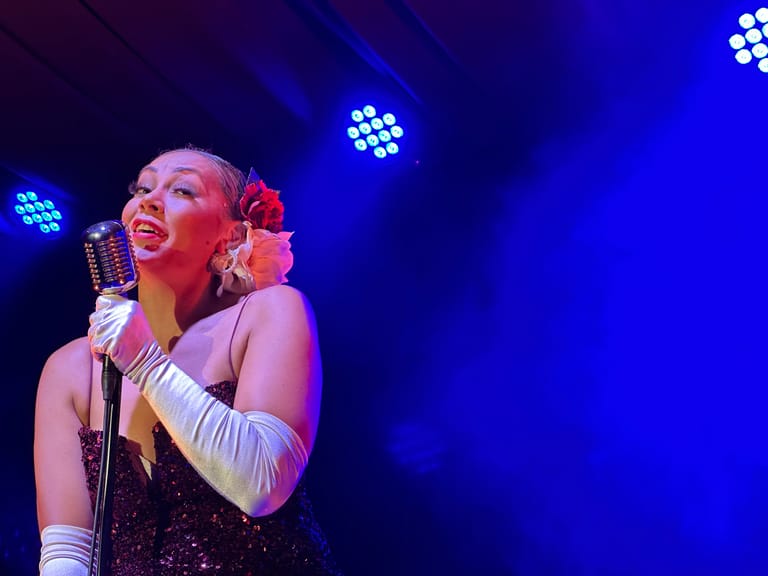Singer in a sequined black dress and white gloves performs at a microphone under blue stage lighting.