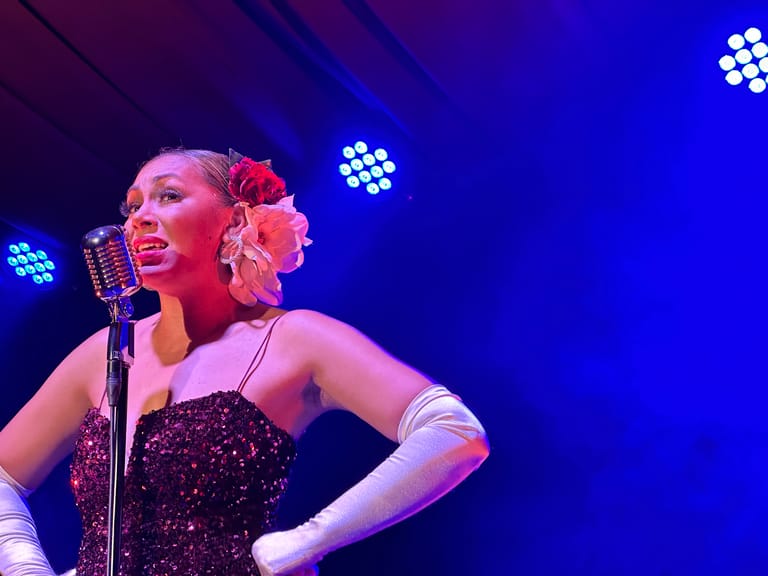 Performer in sequined dress and long gloves singing at vintage microphone under blue stage lights with floral hair accessory.