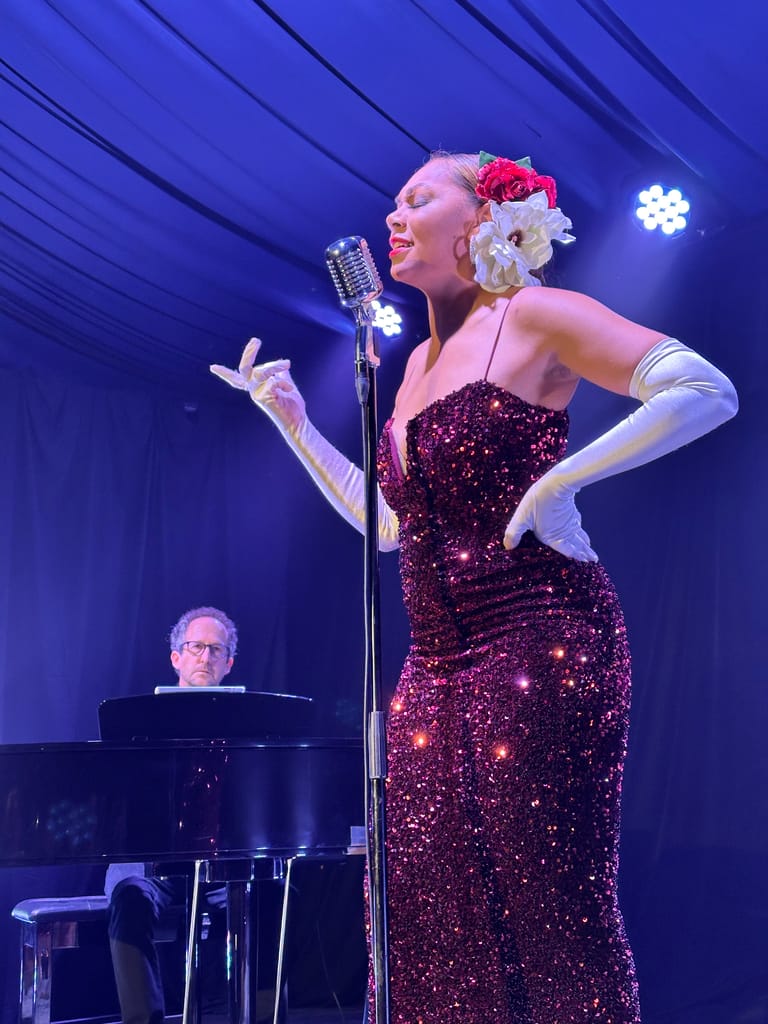 Singer in burgundy sequined gown and floral headpiece performs at vintage microphone with pianist accompanying.