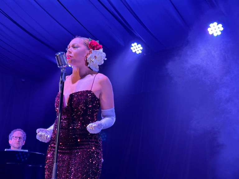 Performer in a sequined burgundy gown and white gloves sings at a microphone beneath blue stage lights.
