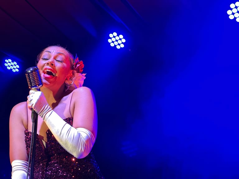 Singer in a sequined dress and long white gloves performs at a microphone under blue stage lighting.
