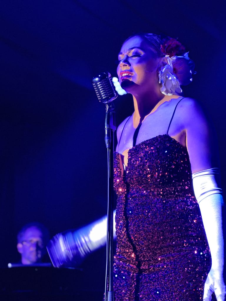 Singer in sequined dress performing at vintage microphone under blue stage lighting, with pianist visible behind.