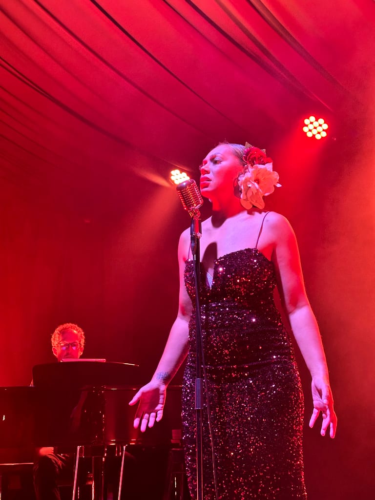 Singer in a sequined black dress performs at a microphone under warm red stage lighting, with a pianist visible at left.