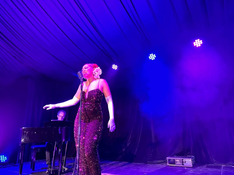 Singer in a sequined gown performs under purple stage lighting with piano accompaniment.