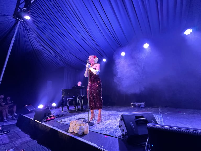 Performer in a burgundy dress singing at a microphone on a stage bathed in blue light, with a piano visible to the side.