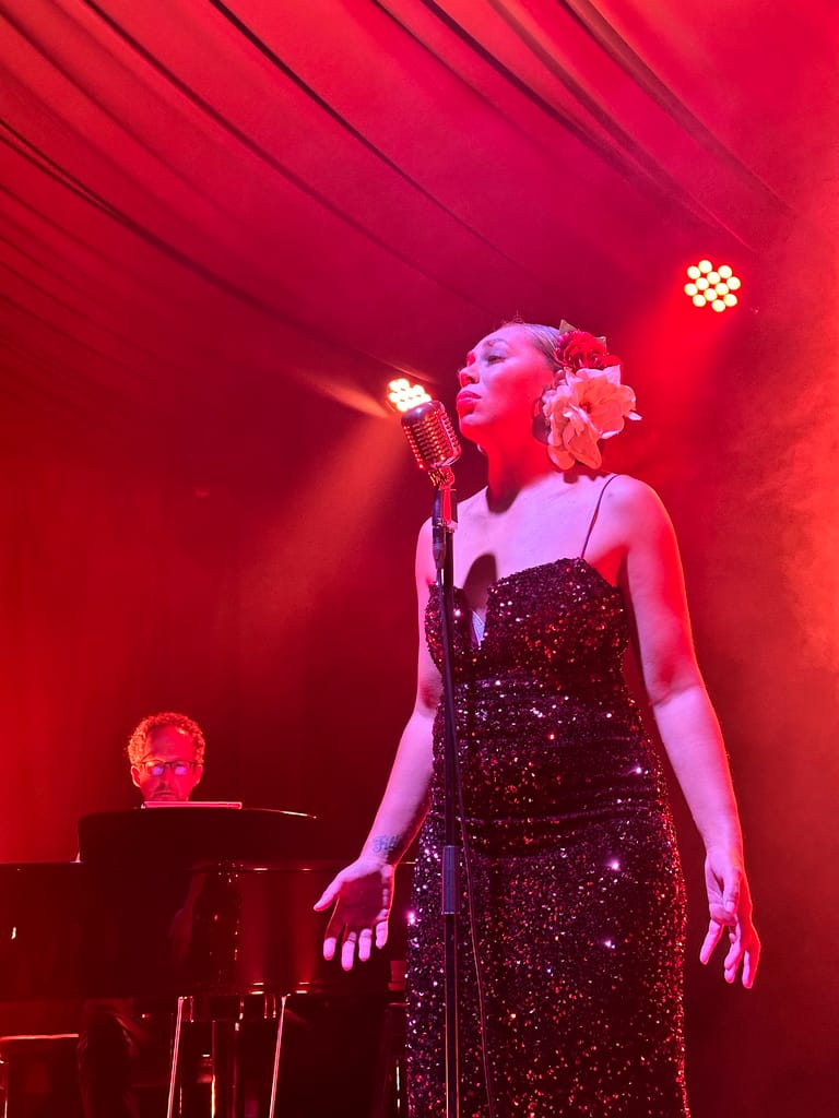 Singer in a sequinned black dress performs at a microphone bathed in red stage lighting.