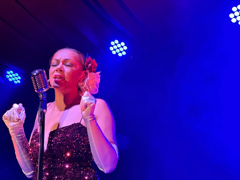Singer in sequined black dress performing at microphone under blue stage lighting.