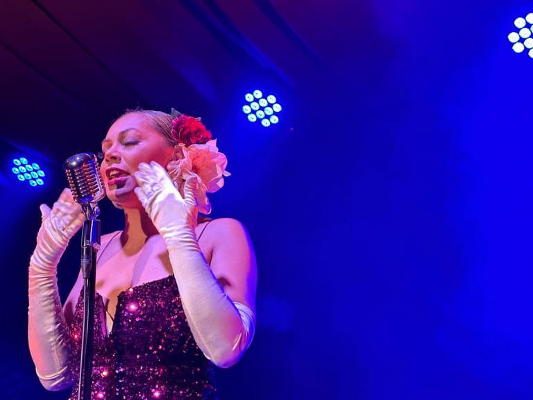 Performer in a sequined dress and long gloves sings at a microphone under blue stage lighting.