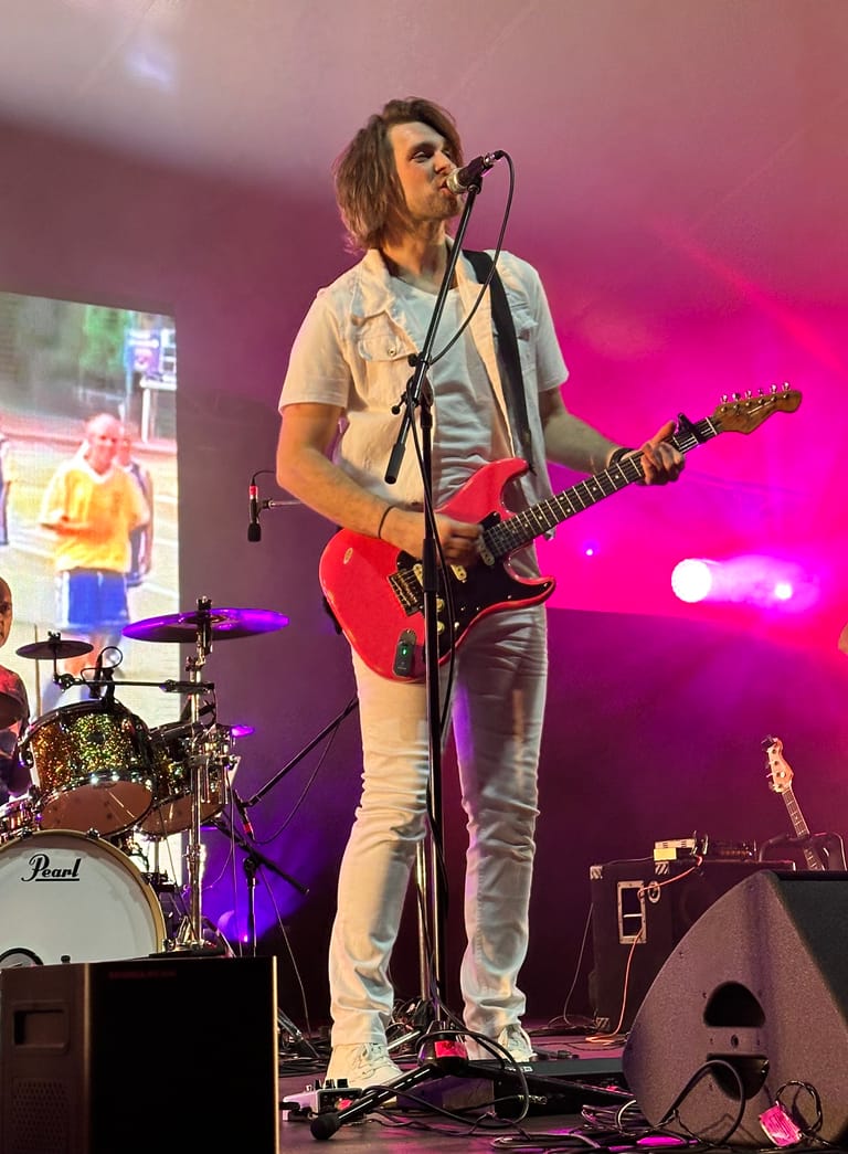 Guitarist in white shirt and red electric guitar singing at microphone under vibrant pink stage lighting.