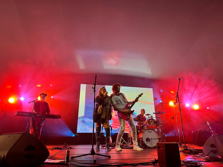 Band performing on stage under pink and red stage lighting with projection screen backdrop.