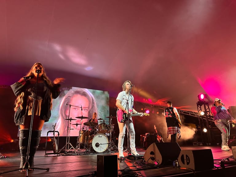 Band performing on stage with pink and purple lighting, featuring a large screen displaying a band member's face.