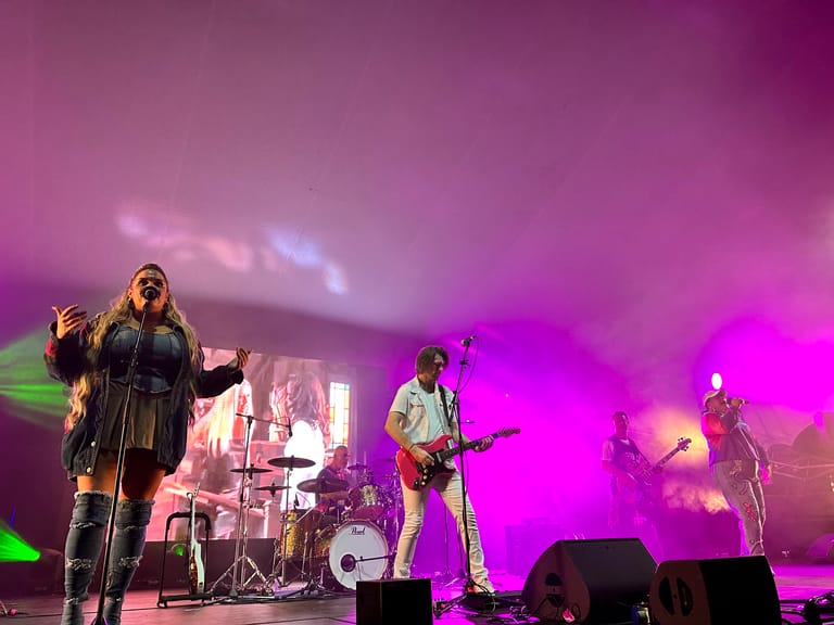 Four musicians perform on stage beneath vibrant magenta and green lighting during a 90s-themed concert production.
