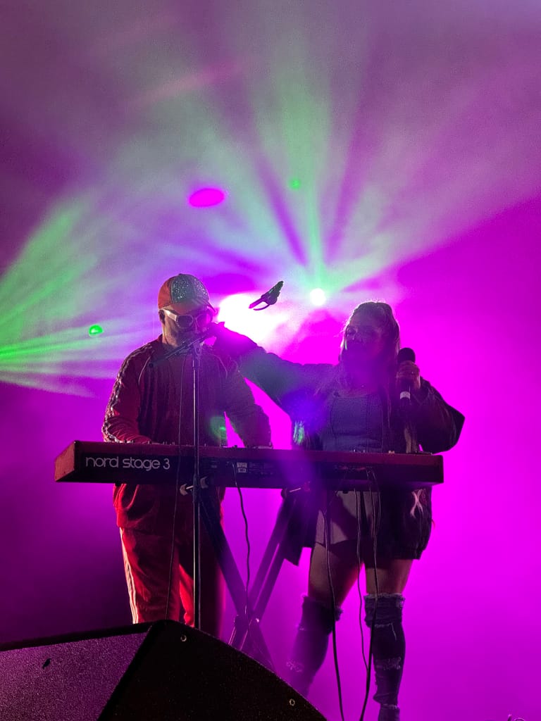 Two musicians perform on stage with synthesizers under vibrant pink and green stage lighting during a 90s-themed show.