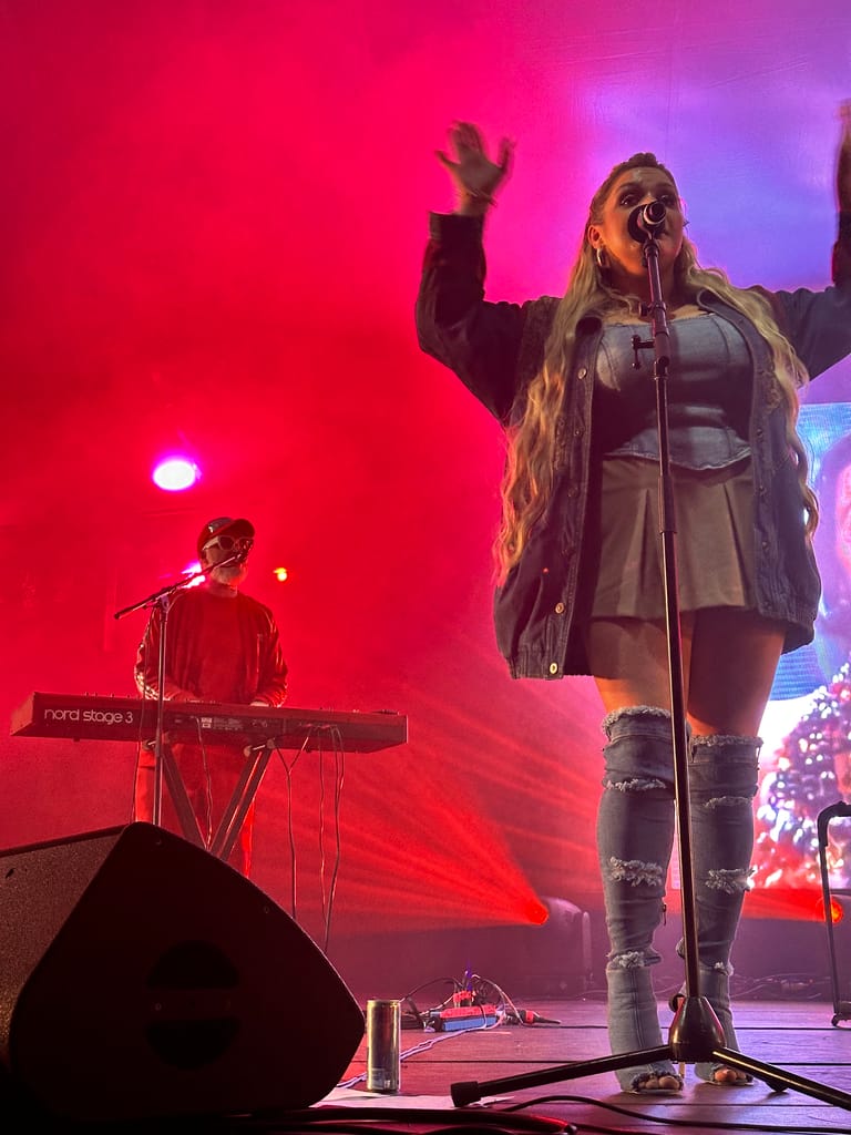 Performer singing at microphone with raised arm whilst keyboardist plays beneath red and purple stage lighting.