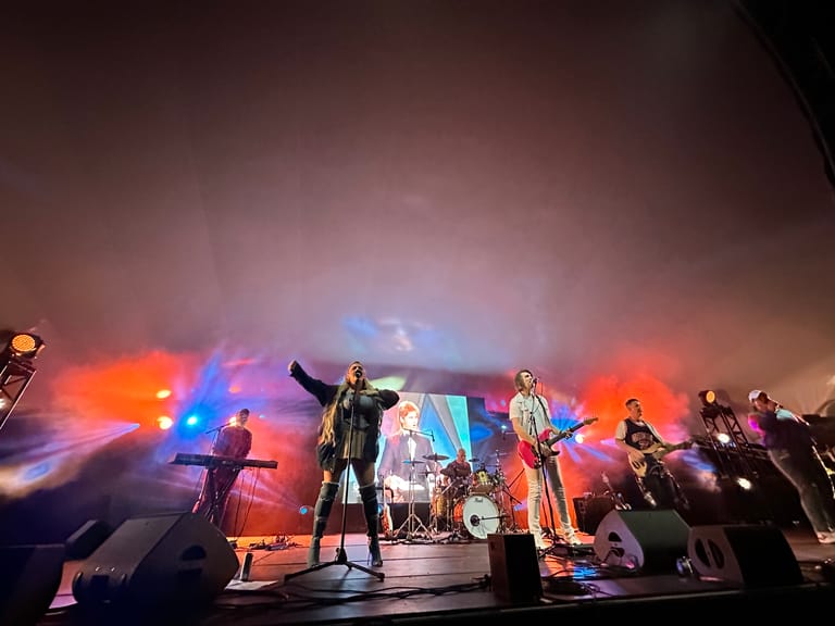 Band performing on stage under colourful orange and blue stage lighting with multiple musicians and instruments visible.