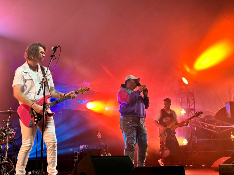 Musicians performing on stage with red and orange stage lighting during a 90s-themed live concert production.
