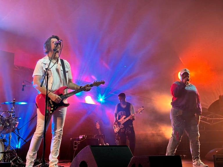 Four musicians perform on stage bathed in vibrant pink, blue and orange stage lighting during a 90s-themed concert.