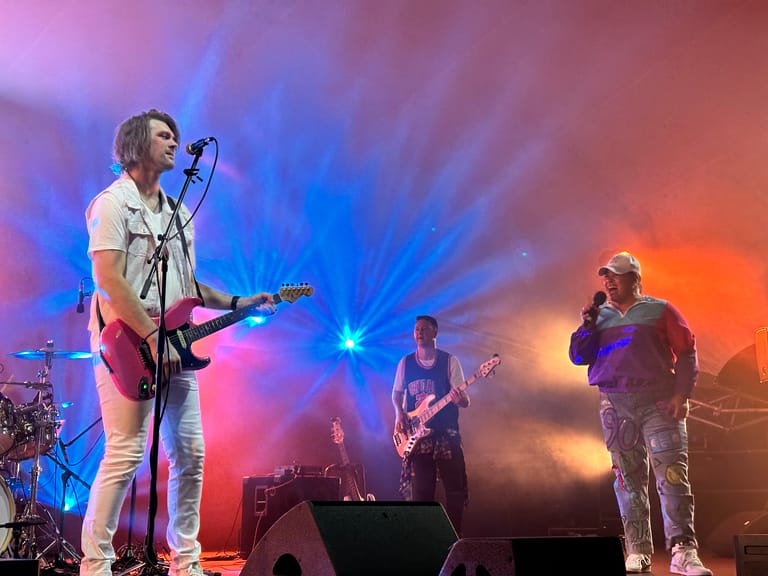 Band performing on stage with colourful blue and orange lighting, lead singer at microphone with guitarist and additional performers visible.