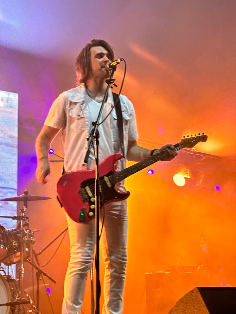 Musician performing with red electric guitar at microphone under warm orange and purple stage lighting.