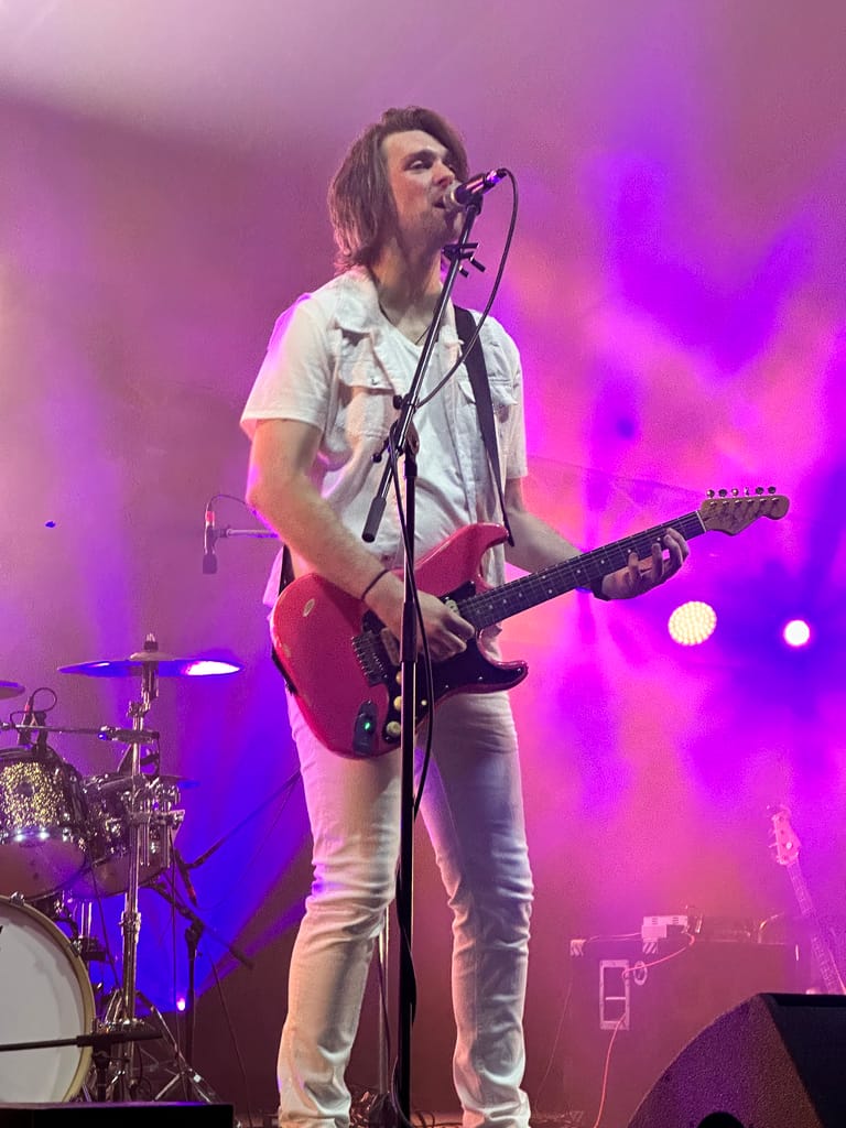 Guitarist and vocalist performing on stage in purple and blue stage lighting during a 90s-themed show.